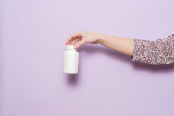 White plastic bottle in female hand in lilac background. Packaging for pills, capsules or supplements.