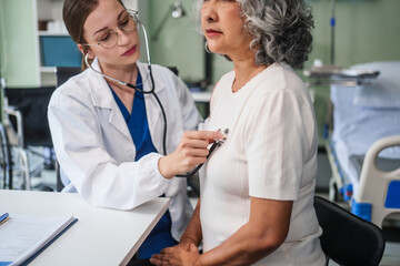 Caucasian woman checks an Asian elderly woman's heartbeat using a stethoscope. They communicate calmly and kindly.