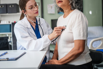Caucasian woman checks an Asian elderly woman's heartbeat using a stethoscope. They communicate calmly and kindly.