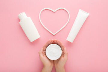 Young adult woman hands holding white cream jar. Bottle, tube and heart shape on pastel pink table background. Care about clean and soft female body skin. Closeup. Point of view shot. Top down view. - 801203148