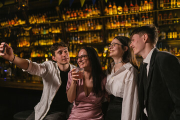 Naklejka premium Men takes out phone and takes photos of close friends after party. Group photo of young people with alcoholic cocktails at bar counter