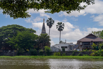 A peaceful riverside scene in Thailand with a historic wat (temple) visible beyond lush greenery and traditional Thai houses, under a clear sky.