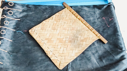 High angle view of a bamboo woven hand fan laid on a blue-colored wooden desk covered by a piece of dark-colored decorative fabric. It commonly used to fan the coals in the charcoal stove when cooking