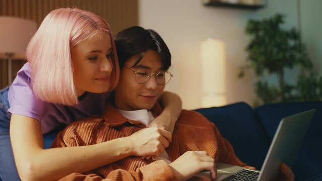 Happy young woman embraces Asian boyfriend booking hotel for vacation on laptop