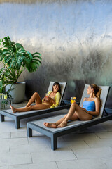 Two women friends are resting on lounge chairs, one of them holding a drink. The scene is relaxed...