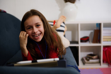 Cheerful little child girl smiles looking at camera, lying on sofa at home, watching cartoon on digital tablet. Little kid have fun on modern gadgets. People. Children, Internet and digital addiction