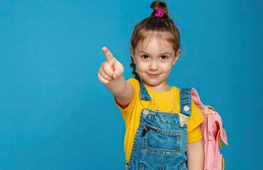 A little girl points her finger forward at the camera on a blue background. Copy space.