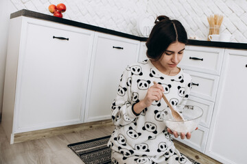 Cheerful woman sitting in bright kitchen mixing dough in a bowl, making homemade cookies, dressed...