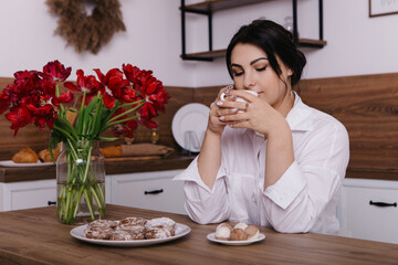 Woman poses with vibrant bouquets of red tulips in vase in kitchen, drink hot tea beverage coffee, eat sweets. Spring International Women`s Day, 8th of March. Holiday, festive, celebration