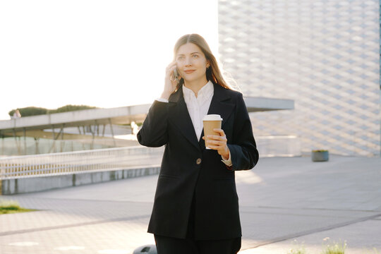 A businesswoman in a black suit walks with a coffee cup in front of office buildings and talking on the phone