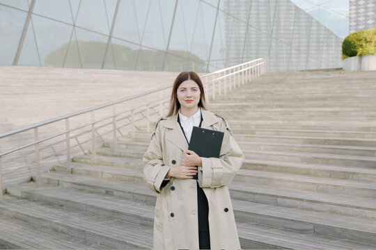Confident young businesswoman in a white shirt and coat with a folder in her hands in front of office buildings