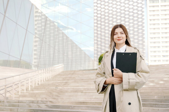 A portrait of businesswoman in white shirt and coat with a folder in her hands in front of office building