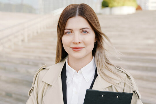 A portrait of businesswoman in white shirt and coat with a folder in her hands in front of office building