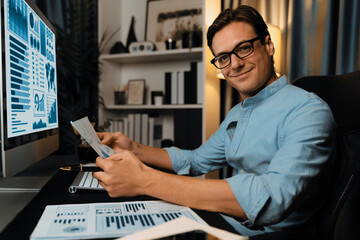 Smart businessman holding paperwork report, looking at camera, profile of remotely working with marketing data report on pc screen, analyzing global target company at modern home office. Pecuniary.