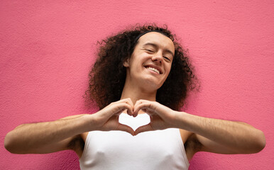 Gay man with curly hair is smiling and making a heart shape with his hands isolated on pink background