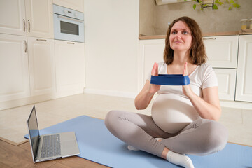 A pregnant woman does sports at home and does exercises with an elastic band for hands stretching, exercises to prepare for childbirth