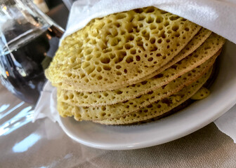 Ployes stacked on a plate covered to stay warm next to a pitcher of maple syrup