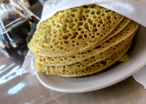 Ployes stacked on a plate covered to stay warm next to a pitcher of maple syrup