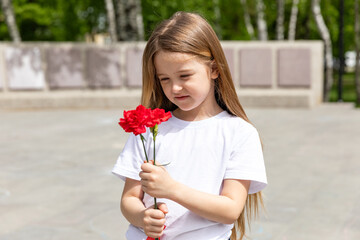 Victory Day concept. Little girl with two red carnations at the Eternal flame near the monument of...