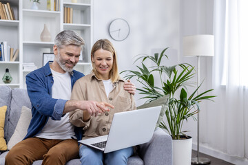 A cheerful middle-aged couple is browsing an online store on a laptop while sitting comfortably on their living room couch, smiling and enjoying a shared shopping experience.