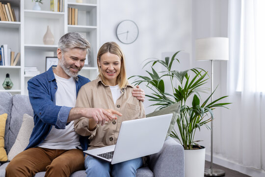 A cheerful middle-aged couple is browsing an online store on a laptop while sitting comfortably on their living room couch, smiling and enjoying a shared shopping experience. - Powered by Adobe