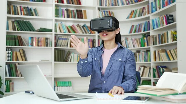 Successful asian female student works using VR glasses in virtual reality simulator sitting in campus library. A young woman uses gestures to control computer programs and flips through virtual pages
