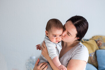 Tired mother sitting on the bed and holding her baby in hands. Trying to calm down baby. Exhausted mother kissing her baby boy. Copy space.