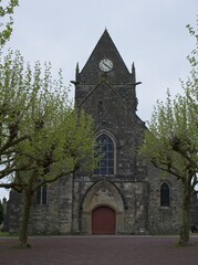 Fototapeta premium Sainte-Mere-Eglise, France - Apr 19, 2024: Sainte-Mere-Eglise church. People walking in Sainte-Mere-Eglise. Streets and buildings. Lifestyle in the urban area. Cloudy spring day. Selective focus