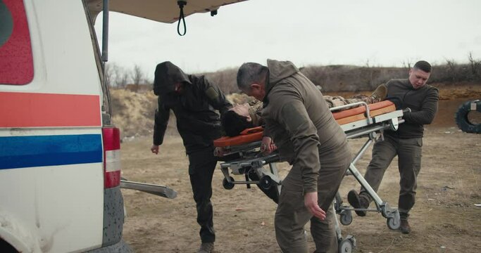 Three confident men and military men in green uniforms put a wounded military man in camouflage uniform on a snare with a doll into an ambulance during a medical emergency exercise at a military