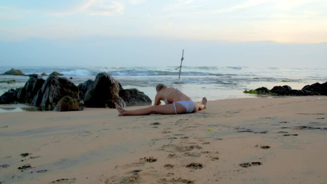 Woman in bikini stretches body performing leg-split on sandy beach. Flexible lady enjoys yoga session by ocean with waves at sunset