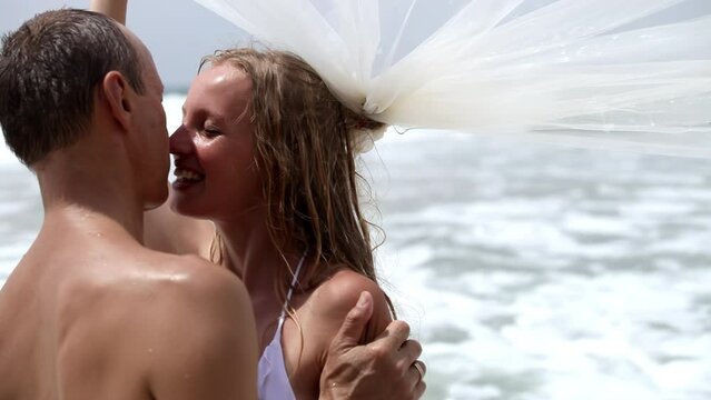 Happy newlyweds share tender kiss standing against ocean waves after ceremony. Bride laughs holding flying veil gracefully in ocean breeze