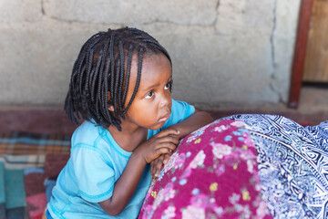 african village, small child girl with braids in front of the house on the porch leaning on her...