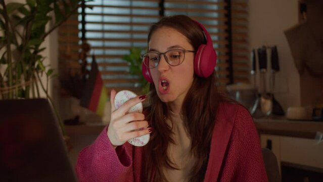 Close up, a young confident brunette girl with glasses in red wireless headphones and a sweater studies a foreign language German and looks at herself with the help of a mirror while pronouncing words
