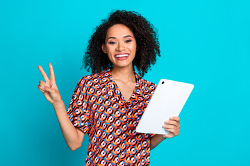 Portrait of cheerful pretty girl with wavy hair wear print shirt hold tablet showing v-sign symbol...