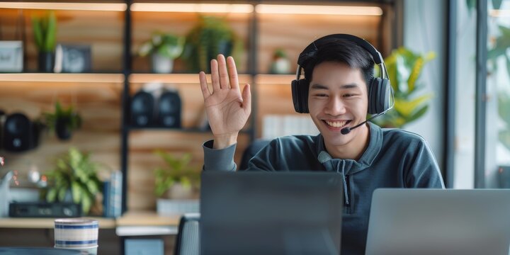 One joyful Asian businessman wearing headset and waving to greet colleagues during laptop-based virtual teleconference meeting in office. Advisor and rep joining global webinar online
