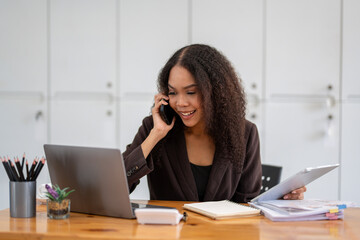 Cheerful African American businesswoman having a phone conversation while working on a laptop in a modern office.
