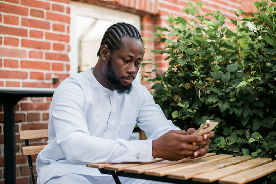 African american man in stylish ethnic dashiki wear viewing photos in social networks on mobile enjoying free time outdoors. Dark-skinned man watching funny video on smartphone and social networks