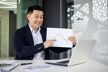 A cheerful Asian businessman in a suit presenting a financial growth chart while discussing business in a modern office settings. He exudes confidence and professionalism.