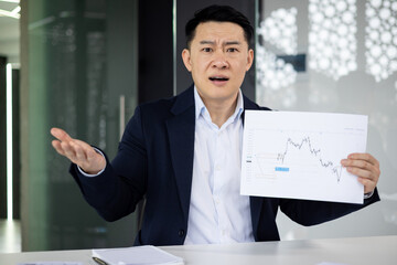 Asian businessman in a suit speaking and gesturing with his hand while holding up a financial graph...