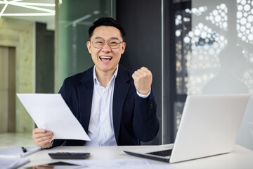 A joyful Asian businessman celebrates a successful deal or achievement at his office desk, showing emotion with a triumphant fist pump.