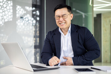 Asian businessman grimacing in pain, clutching his stomach while working at a computer in a modern office setting. Health issues at work.