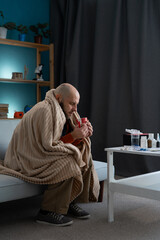 health, cold and people concept. Sad sick man drinking hot tea at home sitting on sofa wrapping in...
