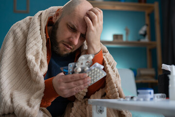 Sick man suffering from fever and headache holds blisters with pills while sitting on the sofa....