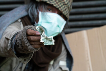 Homeless man wearing a mask and holding out a crumpled dollar bill.
