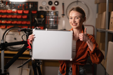 Beautiful Young female mechanic Holding white board With Copy Space For Advertisement, repairman demonstrating white empty billboard with place for text, standing in garage or workshop