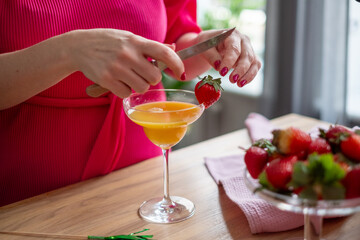 A close up of a hand placing a strawberry on the rim of a glass filled with a golden cocktail, with...