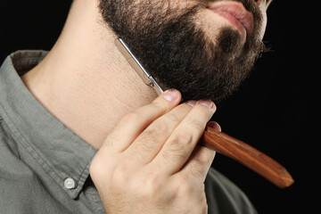 Handsome young man shaving beard with blade on black background, closeup