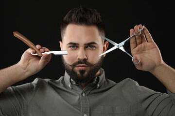 Handsome young man with mustache holding blade and scissors on black background