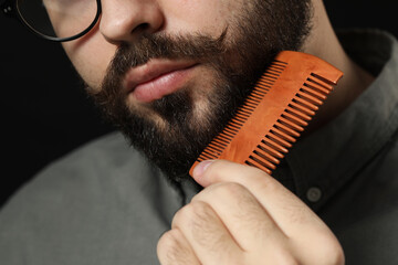 Handsome young man combing beard on black background, closeup