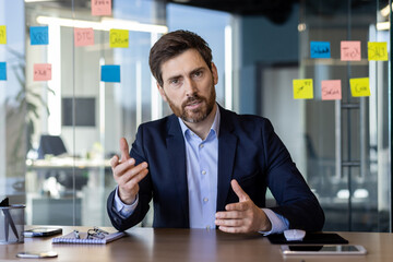 Focused businessman in a suit communicating via video call in a modern office setting, discussing...